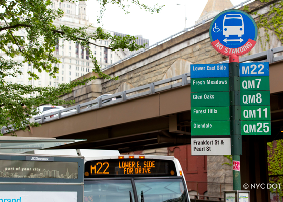 Image of a bus stop pole with the top of a bus and bus shelter in the background.
