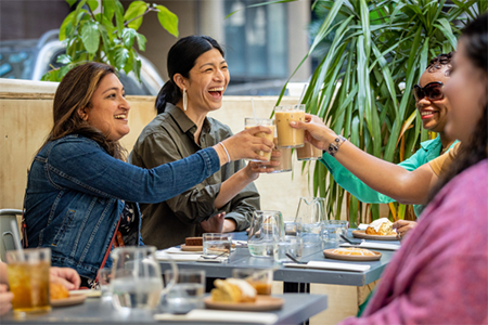 A group of women happily drink coffee while sitting in an outdoor dining area setup along a roadway